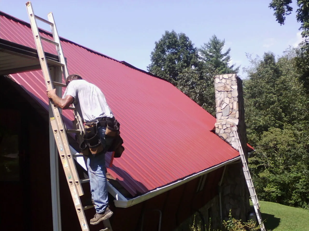 Roofer on a ladder beside a red metal roof and stone chimney — Soffit & Fascia Repair by Roofing Brothers in Holly Bluff